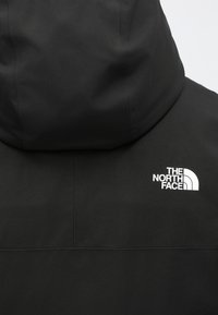 Čierna bunda vyrobená z hladkej tkaniny s kapucňou. Na hornej ľavej strane je biely logo "The North Face". Jednoduchý dizajn.