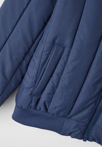 Marineblå polstret jakke med ribbede manchetter, der har en sidelomme og en glat tekstur, der viser et quiltet design med subtile stikninger.