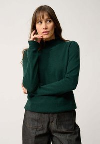 Maglione verde scuro con colletto alto, maniche lunghe e una consistenza morbida, abbinato a pantaloni in denim scuro con tasche visibili e cuciture.