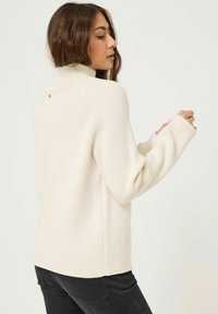 Cremfarbener, gerippter Strickpullover mit hohem Kragen, lockerer Passform, langen Ärmeln und einem kleinen dekorativen Knopf auf der Rückseite.