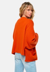 Pull orange surdimensionné avec une coupe large, des manches longues et une poche à l'avant. Fabriqué dans un matériau doux et texturé, associé à un jean bleu clair.