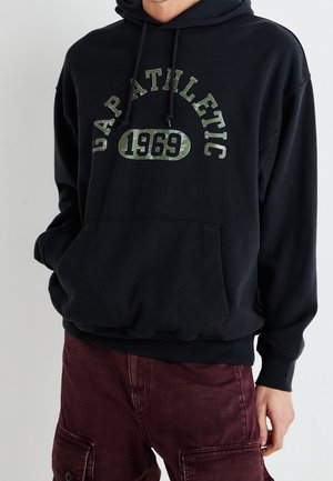 Felpa nera con tasca frontale, caratterizzata da testo verde sbiadito "GAP ATHLETIC 1969" in un carattere in grassetto. Il materiale sembra morbido e accogliente.
