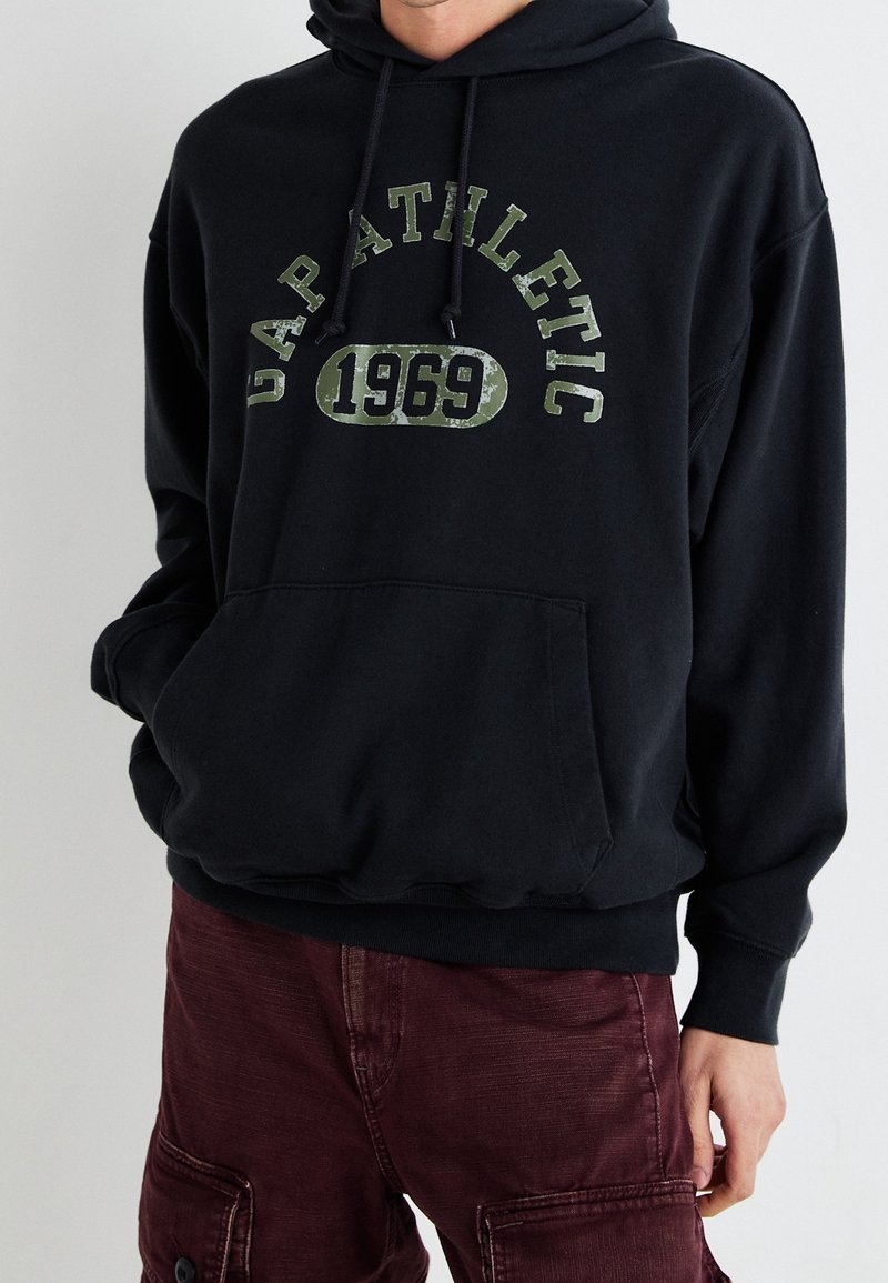 Sudadera negra con bolsillo frontal, que presenta un texto verde desgastado "GAP ATHLETIC 1969" en una fuente en negrita. El material parece suave y acogedor.