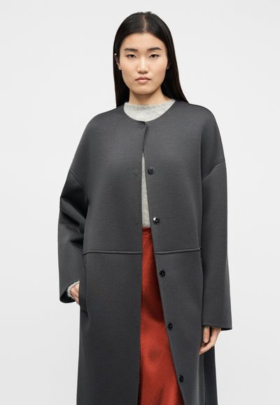 Γυναίκα με μακριά σκούρα μαλλιά φοράει σκούρο γκρι παλτό με κουμπιά πάνω από ανοιχτό γκρι πουλόβερ και κόκκινη φούστα, στέκεται μπροστά σε μονόχρωμο ανοιχτό φόντο.