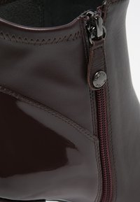 Ciemnobrązowe skórzane botki do kostki z połyskującym wykończeniem, bocznym zamkiem błyskawicznym z czarnymi elementami oraz kontrastującymi bordowymi detalami w postaci przeszyć.