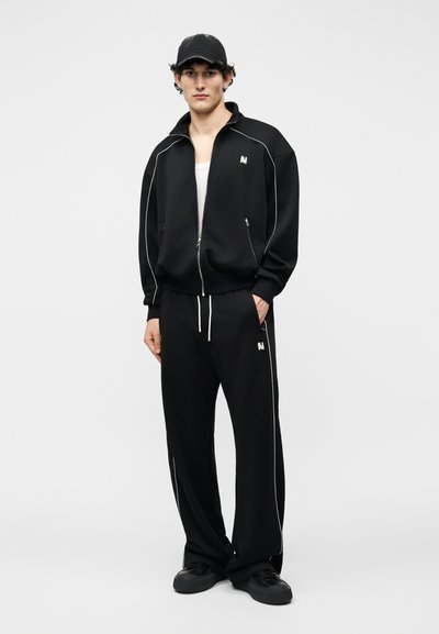 Νέος άνδρας που φοράει μαύρο μπουφάν με φερμουάρ και αντίστοιχο παντελόνι με φαρδιά μπατζάκια και λευκή ρίγα, μαύρο καπέλο, λευκή φανέλα χωρίς μανίκια και μαύρα αθλητικά παπούτσια.