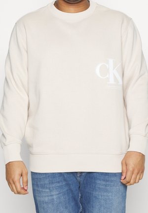 Sudadera beige claro con cuello redondo, puños y dobladillo acanalados. Presenta un logo blanco "ck" en el lado izquierdo del pecho. El material parece suave y liso.