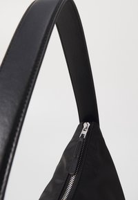 Czarna nylonowa torba z gładkim paskiem ze skóry, wyposażona w srebrny zamek błyskawiczny i kanciasty design. Powierzchnia ma delikatny połysk.