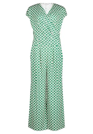 Vrouw in groene en witte jumpsuit met geometrisch patroon, korte mouwen en witte spits toelopende blokhakken, staand tegen een witte achtergrond.