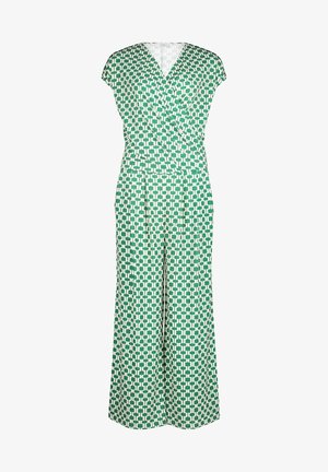 Woman in green and white geometric patterned jumpsuit with short sleeves and white pointed block heels, standing against white background.