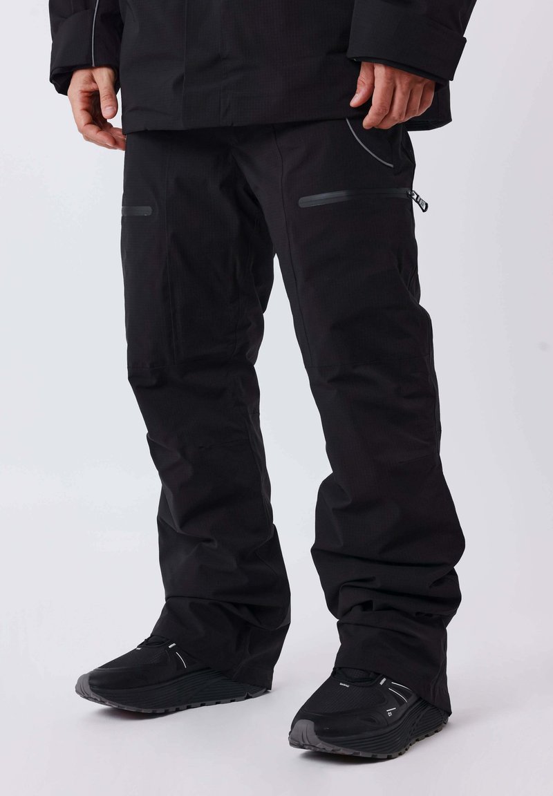 Personne portant un pantalon de plein air noir avec des poches zippées et des chaussures de sport noires, debout sur un fond blanc.