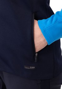 Navyblauwe fleece jack met een ritszak aan de zijkant. Heeft een lichtblauwe mouw en een zwart merketiket op de zak.
