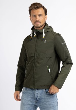 Man die een groene capuchonjas met witte trekkoorden en blauwe spijkerbroek draagt, staand tegen een witte achtergrond.