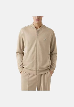 Beige jas met ritssluiting en zakken, geribbelde manchetten en kraag, gedragen met bijpassende beige broek, beide voorzien van kleine driehoekige logo's.