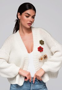 Vit stickad cardigan med stora ärmar, prydd med blommiga applikationer i röd, grön och beige. Bärs över en vit topp och blå jeans.