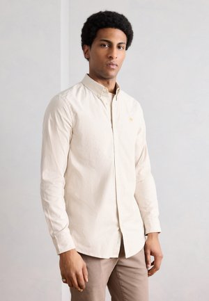 Mand med kort afro-hår iført en beige, stribet skjorte og lysebrune bukser, stående mod en ensfarvet lys væg.
