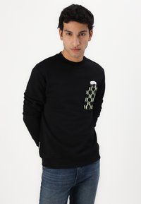 Sort sweatshirt med en frontlomme i et grønt og sort mønster. Lavet af blødt stof, med rund halsudskæring og ribbede manchetter.