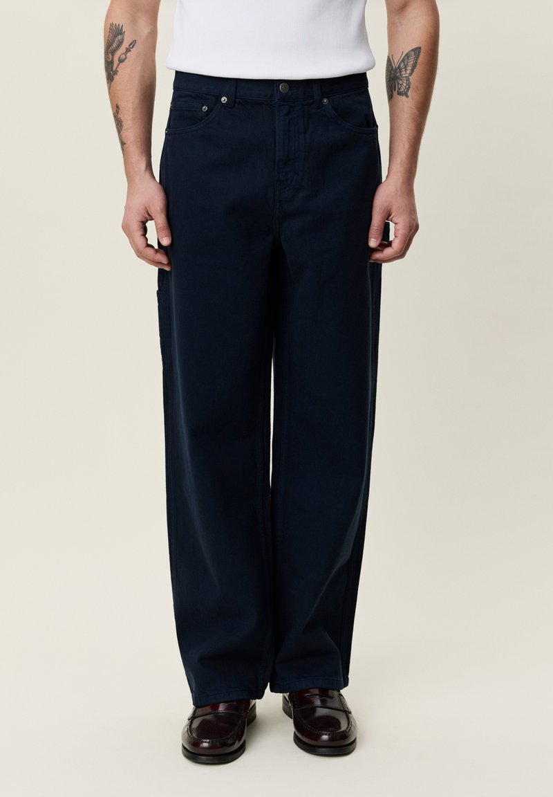 Uomo che indossa pantaloni scuri e larghi, camicia bianca, scarpe eleganti nere e tatuaggi visibili su entrambi gli avambracci su uno sfondo semplice.