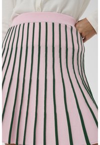 Fustă tricotată roz cu dungi verticale de culoare verde închis, talie lată, purtată cu un top alb cu mâneci lungi, cu mâna sprijinită pe șold.