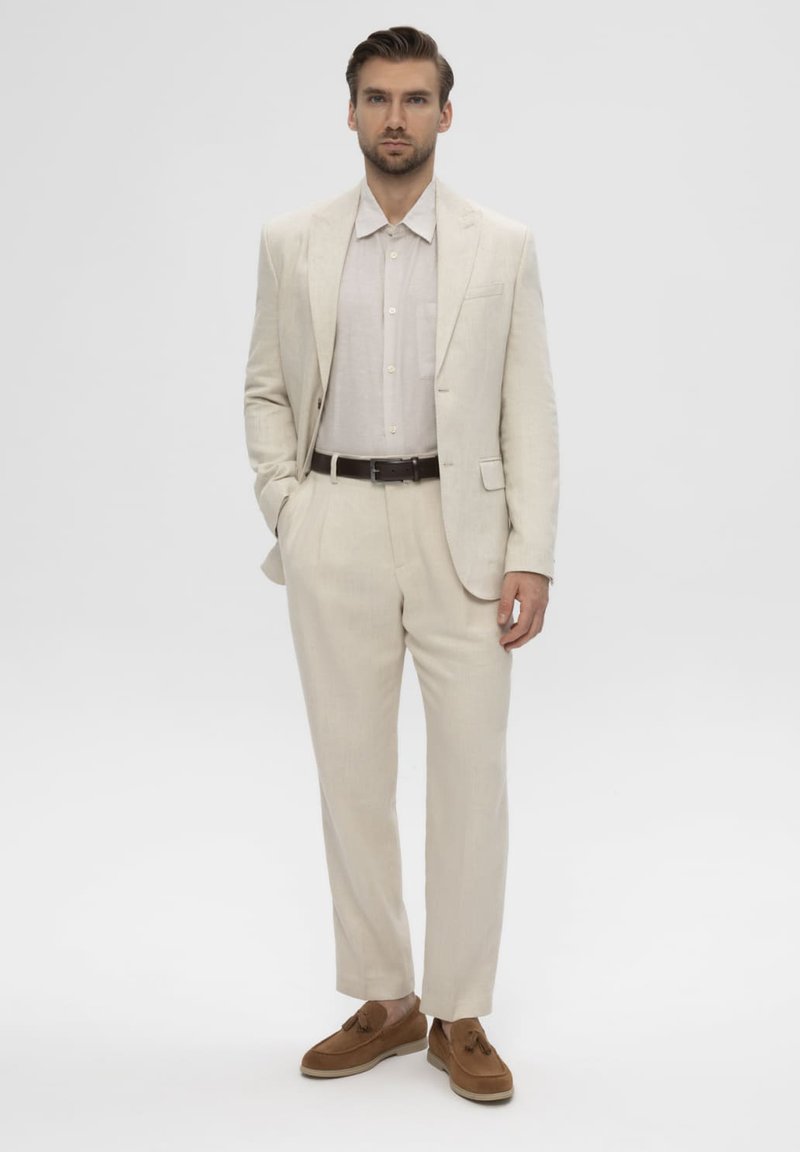 Homme debout portant un costume beige avec une chemise claire, une ceinture sombre et des mocassins à pampilles marron sur un fond clair uni.