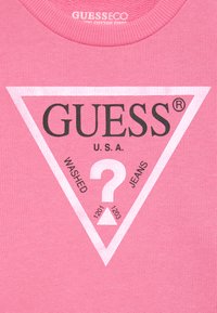 Rosa bomull t-shirt med en stor vit triangelgrafik och svart text. Innehåller texten "GUESS U.S.A." och "? WASHED JEANS" nedanför.