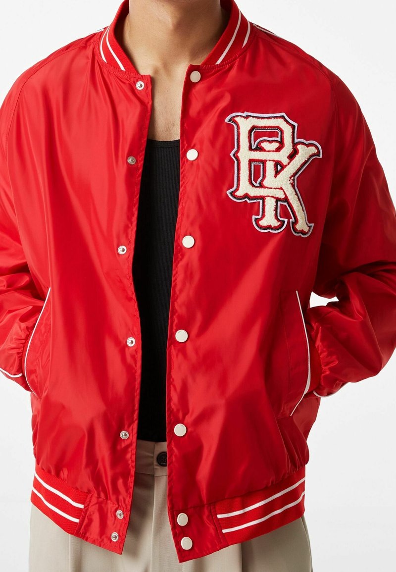 Rode baseballjack gemaakt van glanzend materiaal, met een geribbelde kraag, witte accenten en een geborduurd letterpatch op de borst.