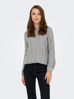 Donna con lunghi capelli castani che indossa un maglione grigio lavorato a treccia e pantaloni neri, in piedi davanti a uno sfondo chiaro uniforme.