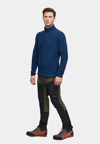 Granatowa polarowa kurtka z wysokim kołnierzem, dwoma bocznymi kieszeniami; zestawiona z czarnymi i oliwkowymi spodniami z kieszeniami cargo oraz butami z pomarańczowymi akcentami.