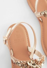 Guldtonade sandaler med en texturerad rem, dekorativa ädelstenar och en slät tan fotbädd, märkt med "FOREVER COMFORT."