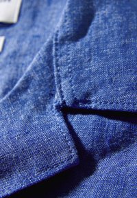 Niebieska tkanina lniana o fakturowanej powierzchni, z kołnierzykiem i detalem przeszycia. Zbliżenie uwypukla kolor i strukturę.