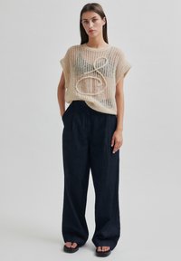 Světle béžový průsvitný úpletový top s žebrovanou texturou a oversize střihem, kombinovaný s tmavě modrými kalhotami širokého střihu, doplněný černými sandály.