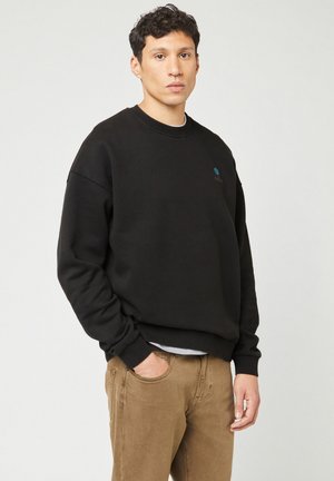 Black sweatshirt made of soft fabric, featuring a crew neck, ribbed cuffs, and a small blue logo on the chest. Worn with beige pants.