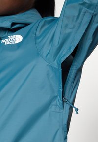 Jachetă albastru deschis cu logo pe piept, prezentând o ziper deschisă pentru ventilație la subsuori și o parte a glugii pe un fundal întunecat.