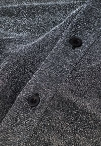Chemise noire boutonnée en tissu texturé et scintillant. Elle présente deux boutons noirs et un subtil détail argenté partout.