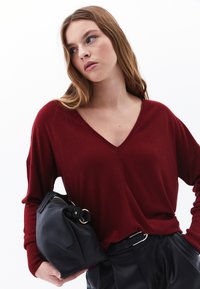 Maglione bordeaux a V, realizzato in tessuto a maglia leggero, con polsini a costine. Borsa a tracolla nera in pelle con dettagli in oro. Pantaloni neri con cintura visibili.