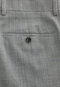 Tessuto grigio con trama, caratterizzato da una tasca posteriore con dettaglio di bottone nero. Il materiale appare leggero con un sottile motivo a quadri.