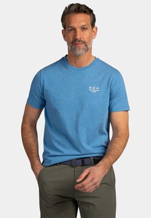 Middelbare man met kort haar en baard draagt een blauw NZA New Zealand T-shirt en olijfgroene broek, staand met hand bij zak.