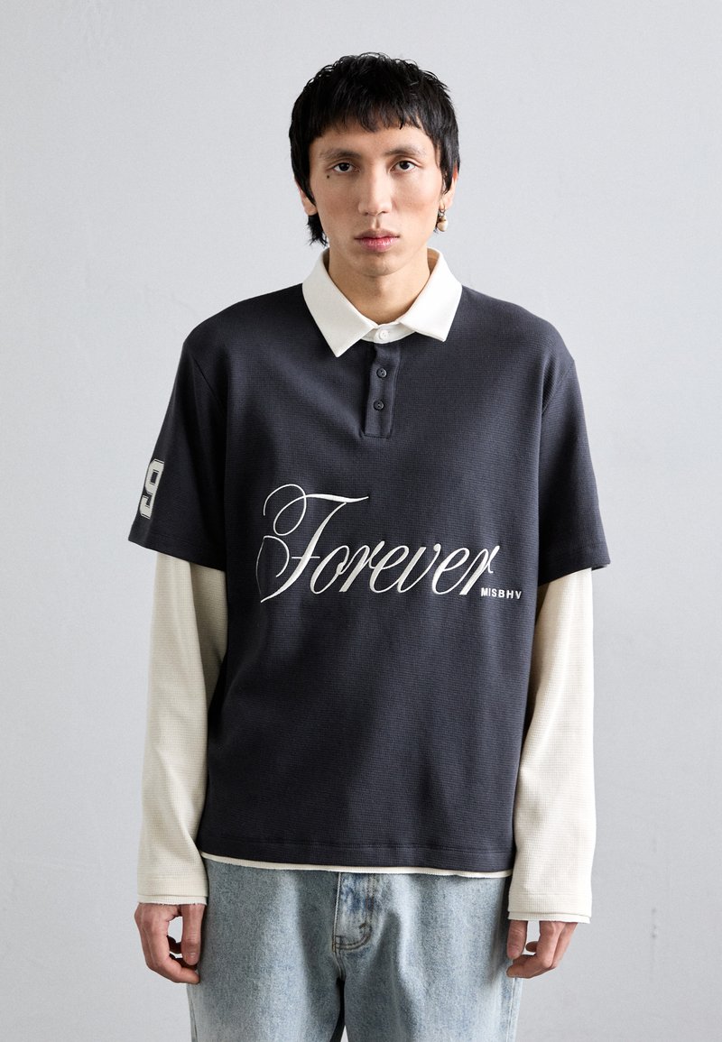 Polo de manga corta gris oscuro con un cuello blanco y la palabra "Forever" en cursiva. Superpuesto sobre una camisa de manga larga clara, combinado con jeans.