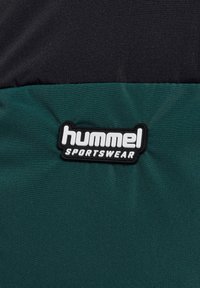 Czarny i zielony materiał sportowy z wypukłym czarno-białym logo „hummel SPORTSWEAR” na fakturowanej zielonej tkaninie.