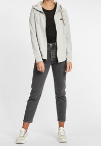 Sweat à capuche gris clair zippé avec un motif tacheté, cordons ajustables et poche kangourou, assorti à un jean noir et des baskets beiges.