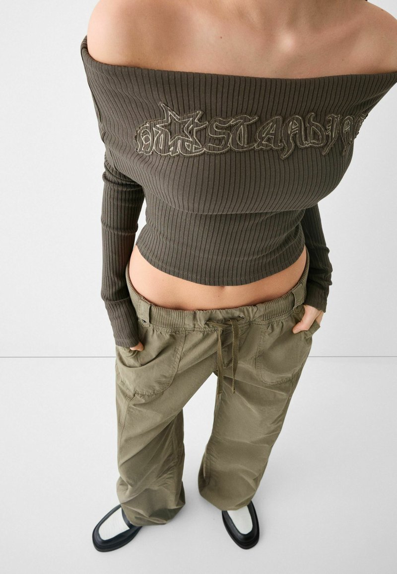 Gerippter, schulterfreier Oliv-Grün Crop Top mit dekorativen Nähten; kombiniert mit lockeren khakifarbenen Cargo-Hosen und weißen Slippern mit schwarzer Sohle.