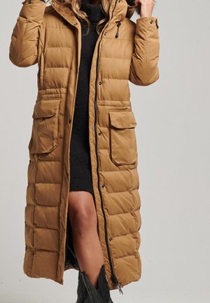 Femme portant un long manteau matelassé beige avec de grandes poches par-dessus une robe noire et des bottines noires, ajustant la capuche du manteau.