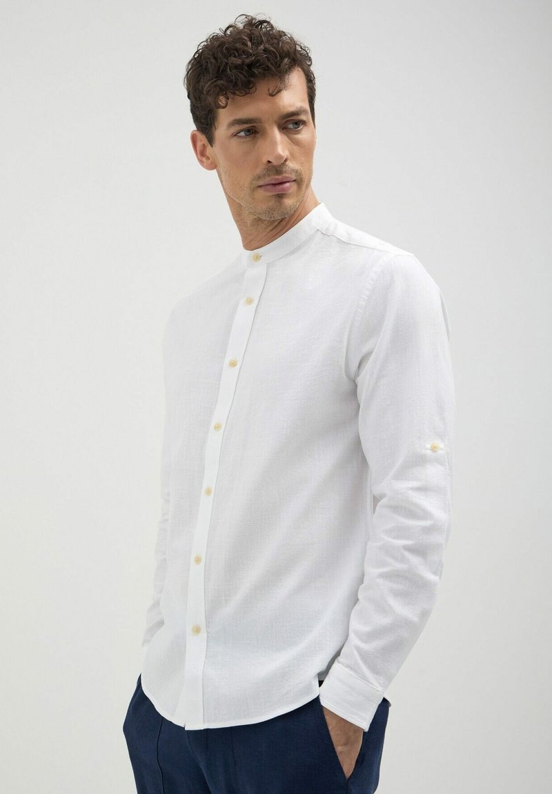 Chemise blanche à manches longues avec col mandarin, présentant des boutons beige clair, un tissu texturé et des poignets retroussés pour un ajustement personnalisable.