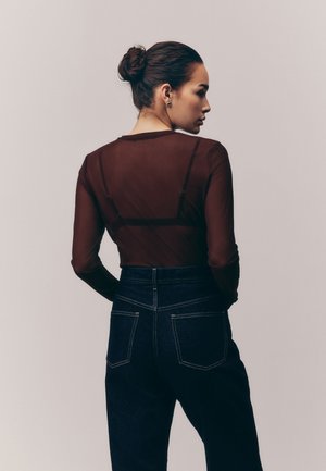 Femme avec les cheveux attachés en chignon, portant un haut transparent sombre à manches longues et un jean en denim foncé taille haute, vue de profil sur un fond uni.