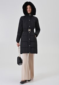 Zwarte donzen jas met een capuchon gevoerd met bont, een tailleband en drukknoopsluiting. Gedragen over beige uitlopende broek, met een zwarte handtas.