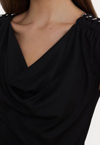 Svart draperad topp i mjukt tyg, med V-ringning, rynkade axlar och metalliska detaljer vid axelsömmarna.