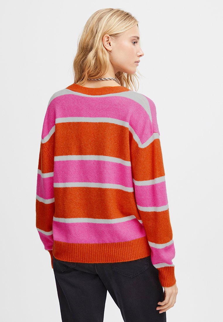 Kobieta z blond włosami, stojąca bokiem, ubrana w sweter w paski o poziomych pasach w kolorze pomarańczowym, różowym i beżowym oraz czarne spodnie.