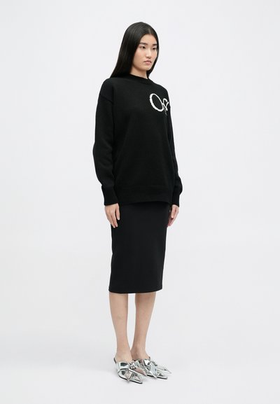 Maglione nero a maglia con polsini e scollatura a coste, impreziosito da un logo ricamato bianco. Abbinato a una gonna nera a tubo e mule metallici.