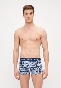 Férfi szűkített boxer alsónemű, sötétkék derékkal, fehér és világoskék "HUGO" logóval, amely ismétlődik a mintás anyagon. Puha, rugalmas anyag.