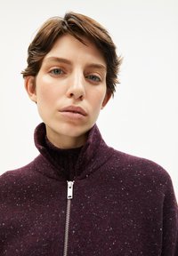 Bordeauxfarbener Reißverschluss-Pullover mit hohem Kragen, strukturiertem Stoff mit helleren Sprenkeln und silbernem Reißverschlussdetail. Nahaufnahme des Kleidungsstücks.